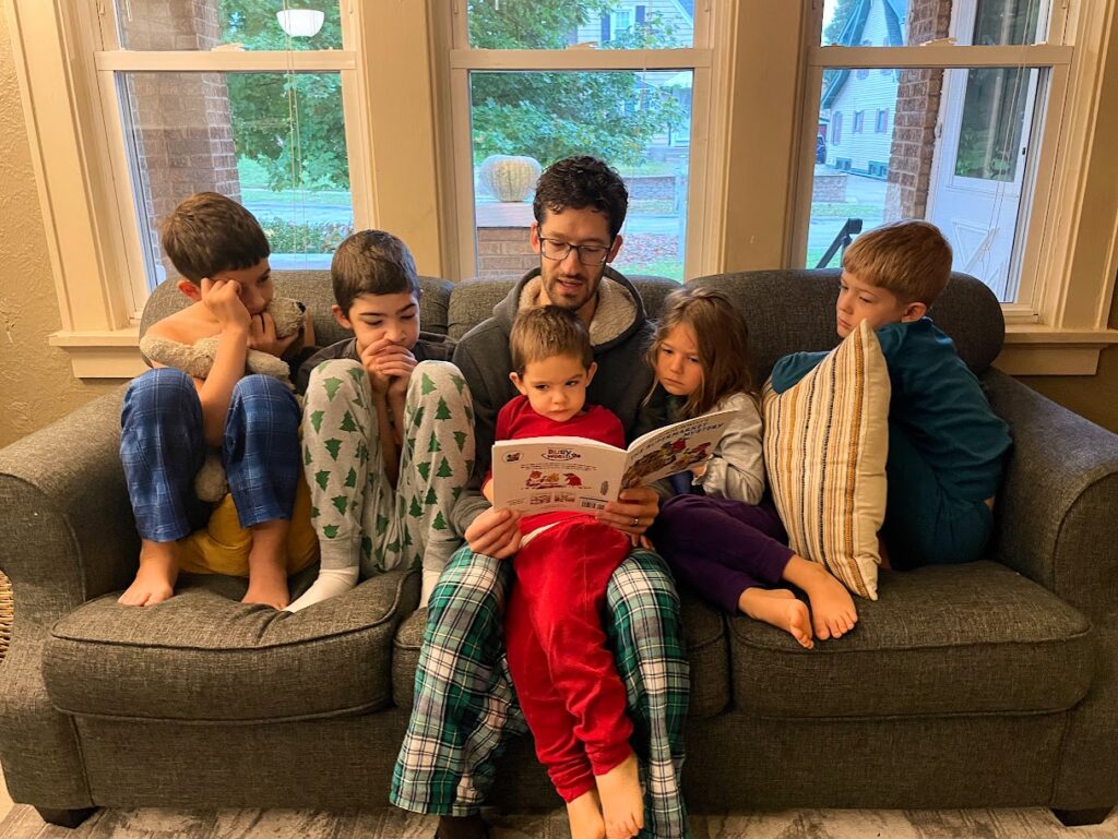 a dad sitting on a couch reading a book to five homeschool children