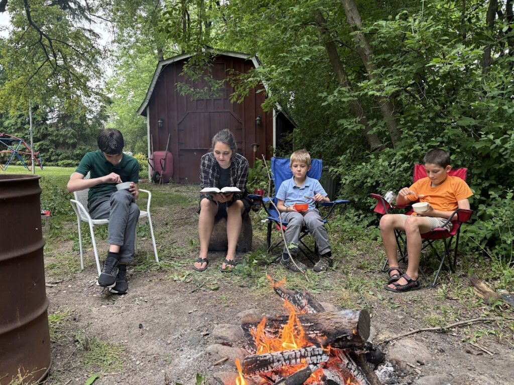 homeschool mom reading aloud to three boys while sitting outside near a campfire
